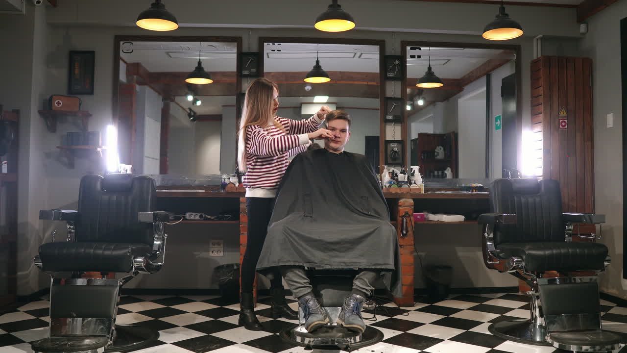 Interior shot of working process in modern barbershop. Side view portrait of attractive young man getting trendy haircut. Male hairdresser serving client, making haircut using metal scissors and comb.