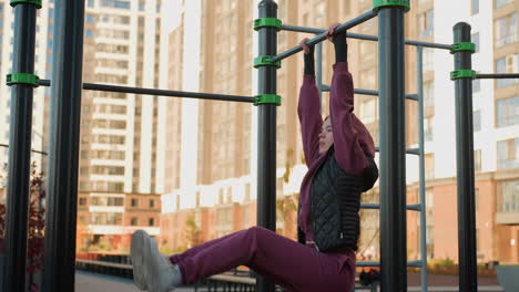 side view of outdoor trainer gripping pull up bars with hands while lifting legs in urban park workout session, focus on core strength, balance, endurance and technique during body weight exercise