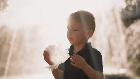 blond little boy eats piece of cotton candy against fountain