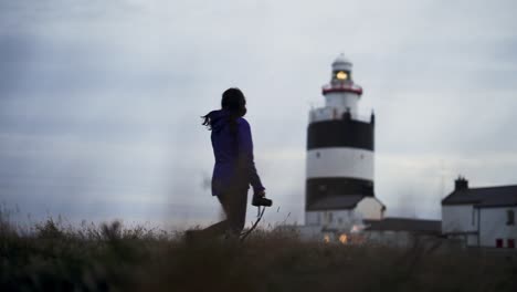 Young-female-photographer-walking-toward-lighthouse-under-overcast-sky