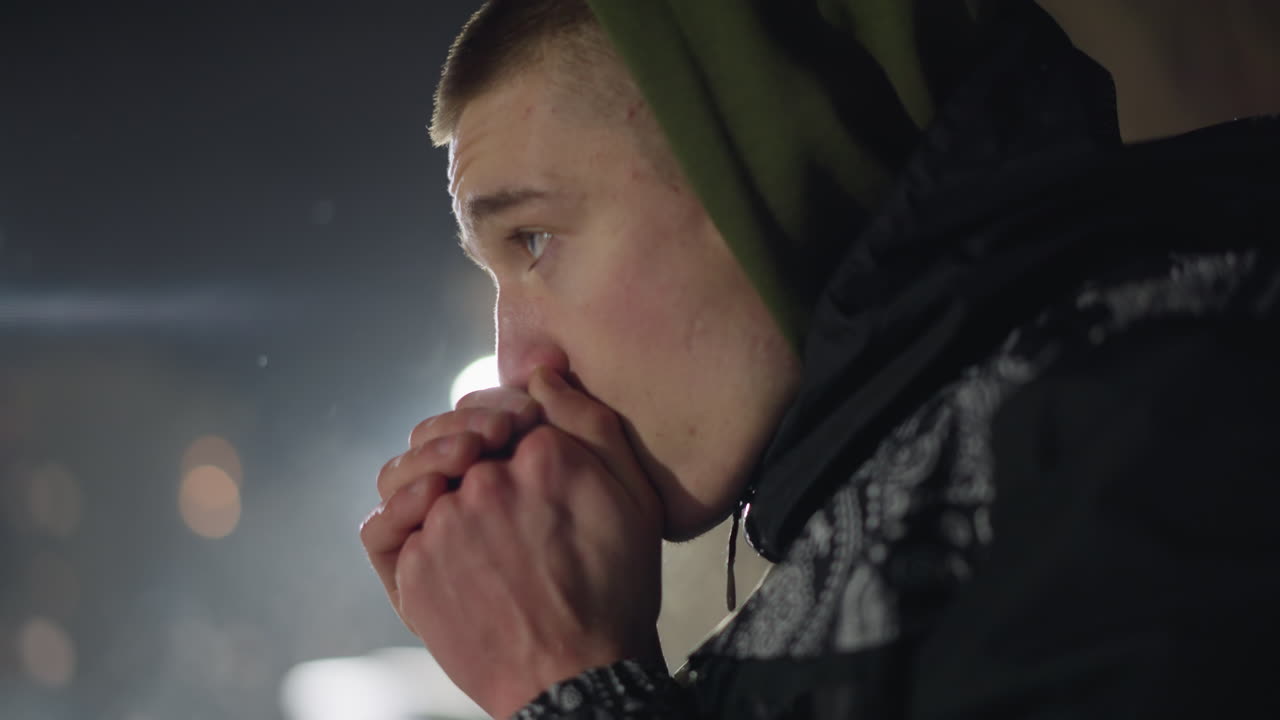 Young man feeling cold during winter blows air into hands to stay warm, visible mist from breath in cold air, distant light illuminating scene with soft bokeh and dark outdoor background