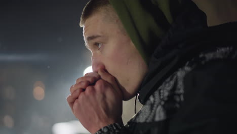 young man feeling cold during winter blows air into hands to stay warm, visible mist from breath in cold air, distant light illuminating scene with soft bokeh and dark outdoor background