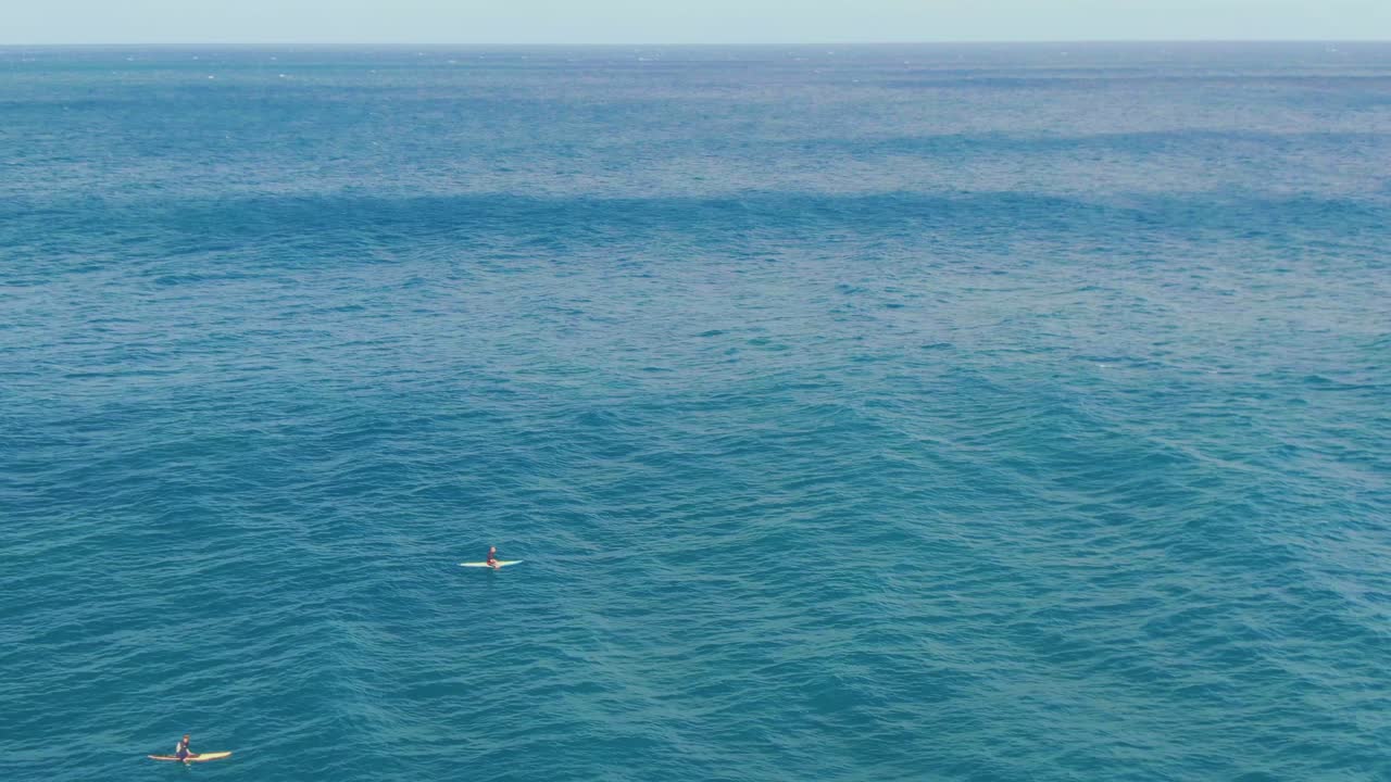 Surfers waiting on their board for big wave to ride, paddling slowly on deep blue ocean near shore of Hawaiian island