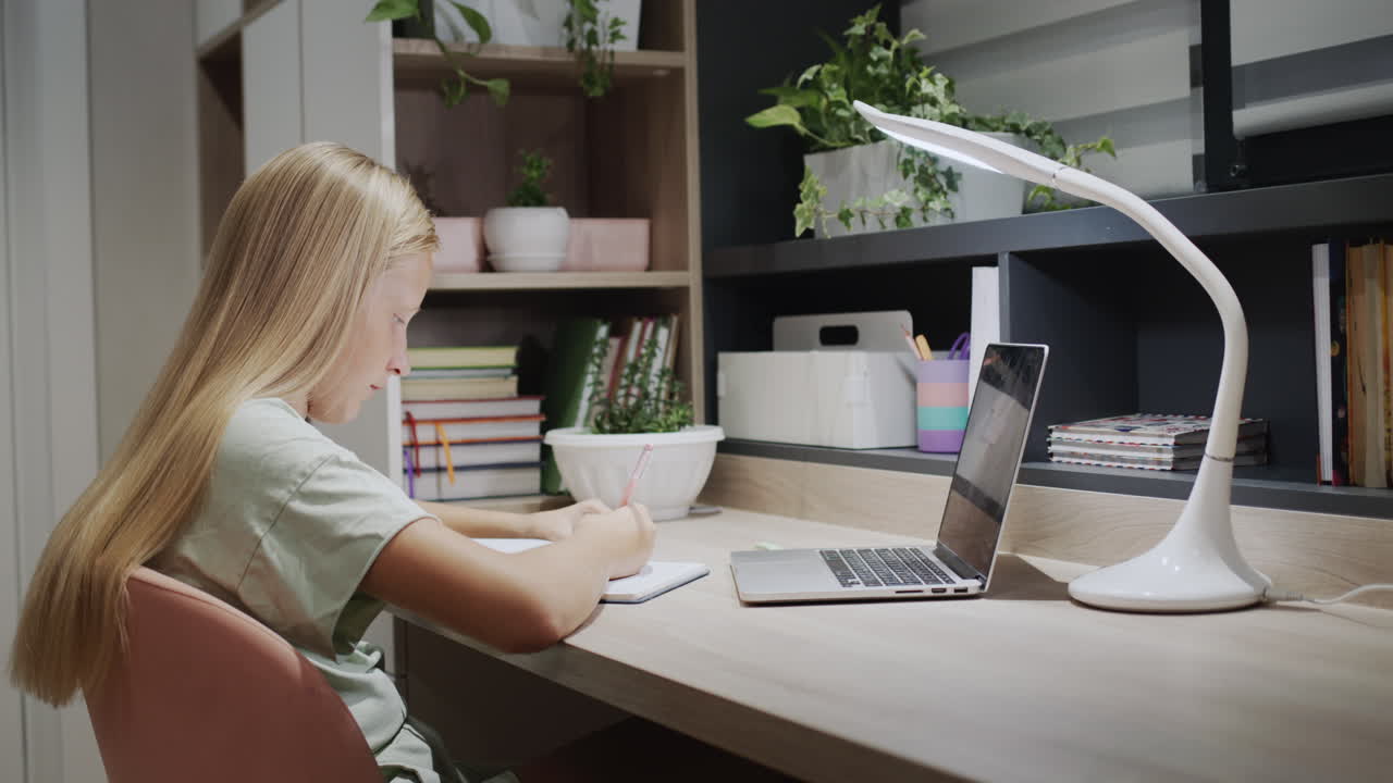 un niño con un hermoso cabello largo hace la tarea en casa, se sienta en una mesa cerca de una computadora portátil