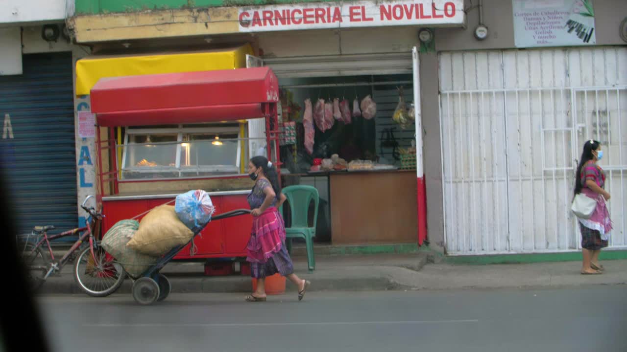 Slow motion street view woman in traditional attire pushing hard cart loaded with huge bags, wearing face mask, Zone 18 Guatemala City - Handheld