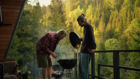 un tipo rubio con una camisa a cuadros roja hace salchichas en la parrilla, y su amigo, un tipo moreno, te dice cómo hacerlo bien. amigos en un picnic con vistas al bosque de coníferas y las montañas