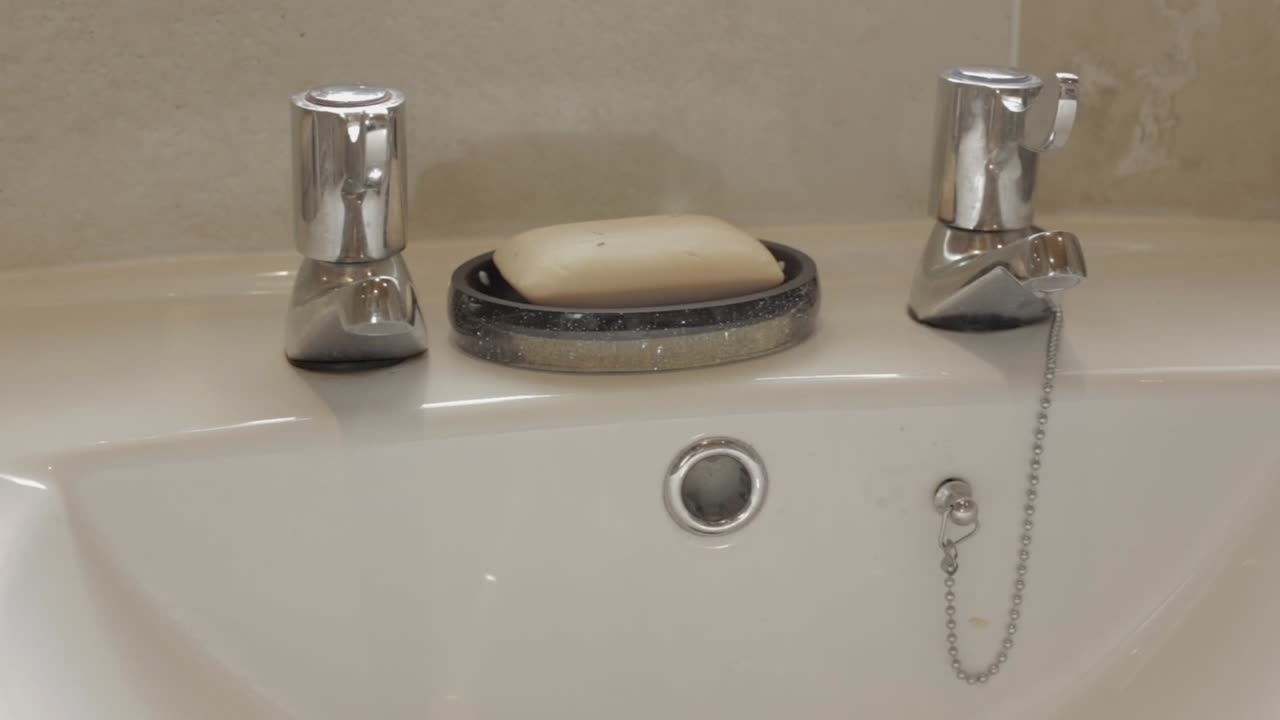 Close up pan shot of sink wash basin with taps and soap against tiled wall