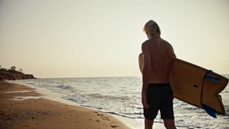 Shooting-on-the-site,-a-blond-man-with-a-bare-torso-in-black-shorts-carries-a-yellow-surfboard-and-walks-along-the-orange-sandy-seashore-in-the-morning-at-Sunrise