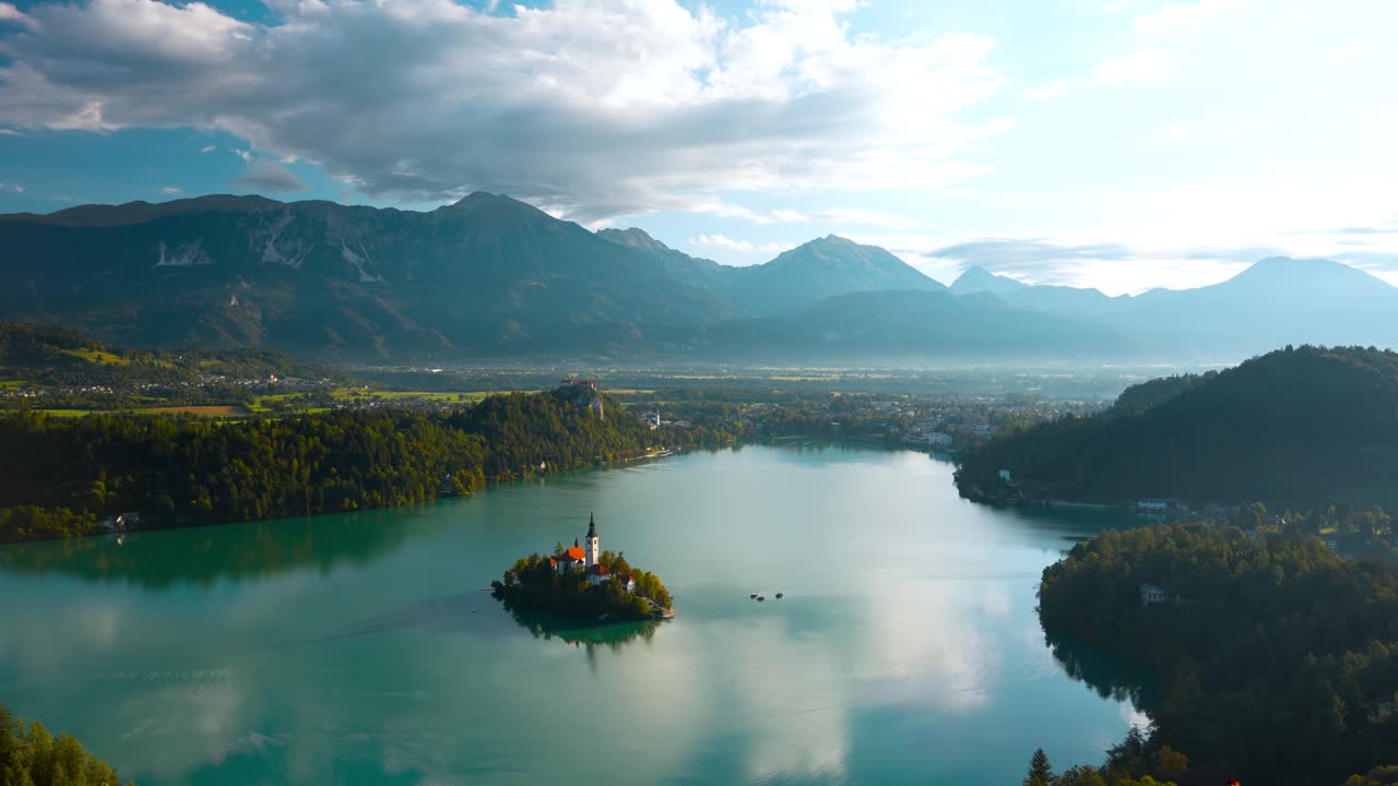 Il lago di Bled circonda la sua chiesa sull'isola sotto le cime alpine e le nuvole, ripresa panoramica fluida da destra, campo largo