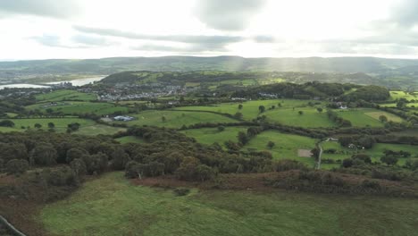 Sunbeams-moving-across-overcast-British-agricultural-rural-village-countryside-morning-aerial-view-rise