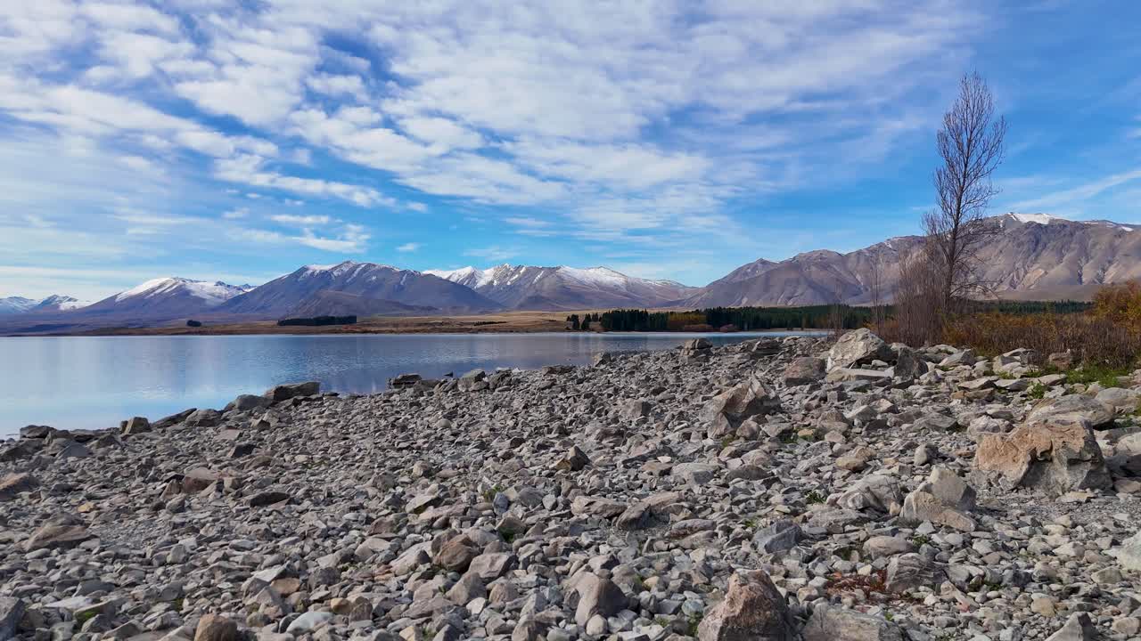 Drone footage captures Lake Tekapo's rocky shoreline with distant snow-capped mountains under a vibrant blue sky