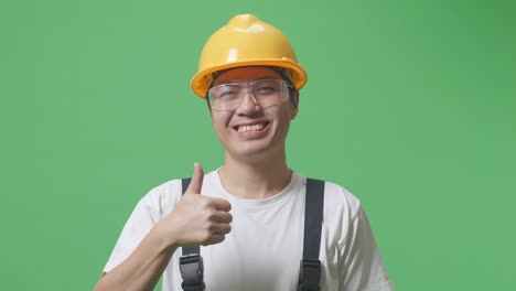 close up of asian man worker wearing goggles and safety helmet smiling and showing thumbs up gesture while standing in the green screen background studio