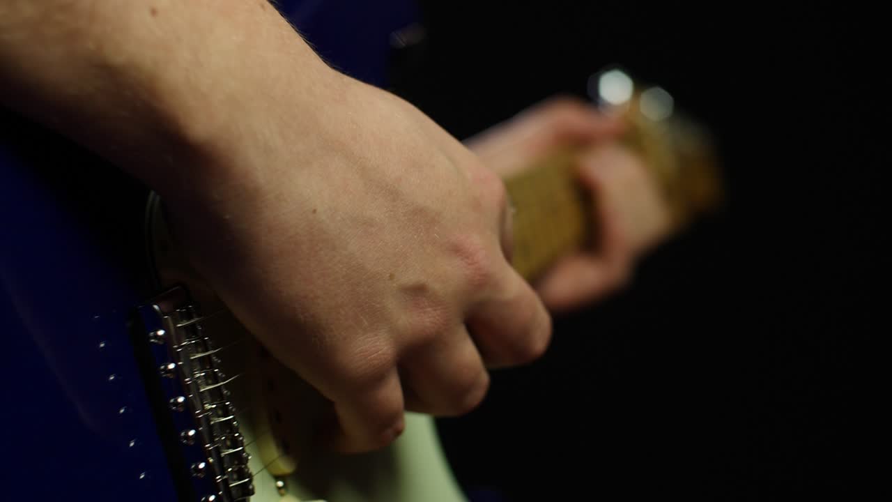Slow motion close up of an electric stratocaster guitar and guitarists hands while playing notes. Smooth slow motion with gimbal backwards movement out of focus, dark background. Shot in 4K at 120fps