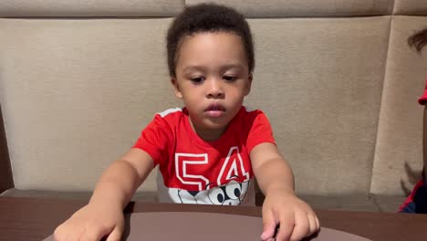 Lindo-Y-Exótico-Niño-Negro-De-Dos-Años-Esperando-Impaciente-Su-Comida-En-Un-Restaurante,-Vestido-Con-Una-Camisa-Roja,-Sentado-Junto-A-Mamá