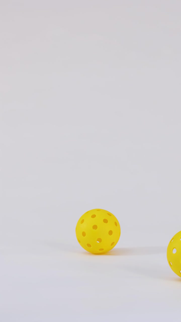 Three yellow plastic pickleball balls fall, bounce, and roll across a clean white studio surface