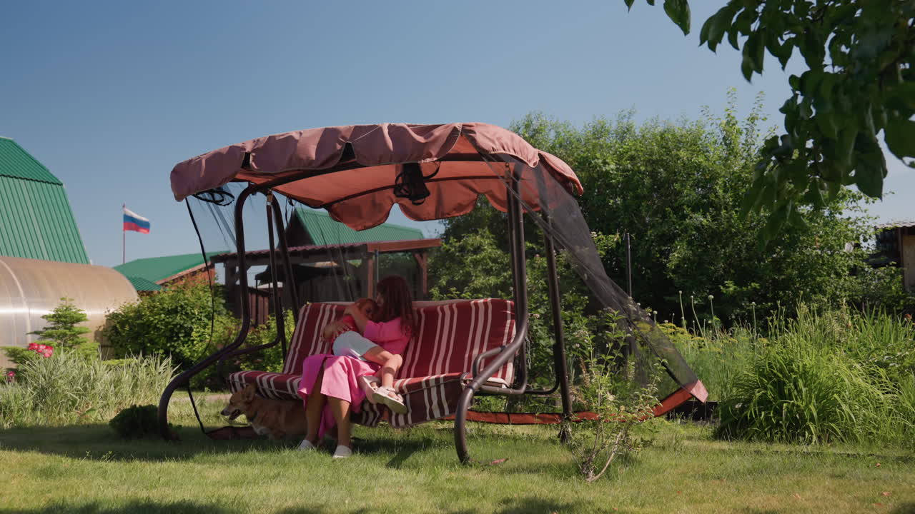 Woman Sitting Peacefully In Garden, Serene Woman In Pink Enjoying Quiet Afternoon Outside, Woman Dressed In Rose Attire Reads Calmly On Garden Swing With Shaded Canopy And Vibrant Surroundings