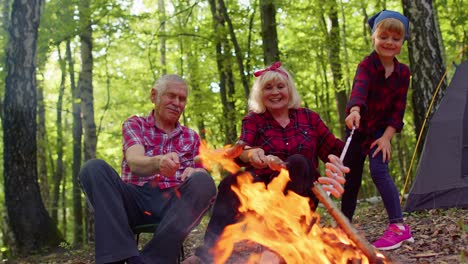 Happy-senior-man-woman-with-grandchild-talking-cooking-frying-sausages-over-campfire-in-evening-wood