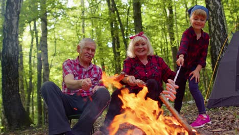 happy senior man woman with grandchild talking cooking frying sausages over campfire in evening wood