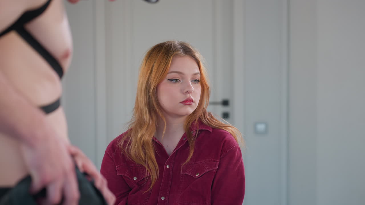 young woman seated in red corduroy shirt watches half dressed male stripper in harness fix trousers then averts gaze before looking back, showing mix of boredom curiosity inside mirrored studio