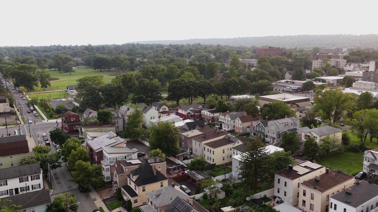 Fitta fila di case coloniali con tetto a capanna costeggia una strada alberata a Orange, New Jersey; auto parcheggiate e campi da gioco verdi in lontananza si estendono oltre i tetti sfocati della città nelle vicinanze. Ripresa aerea con drone in ascesa.