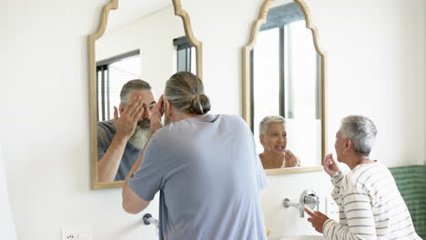 senior couple in bathroom, man grooming beard while woman applying skincare