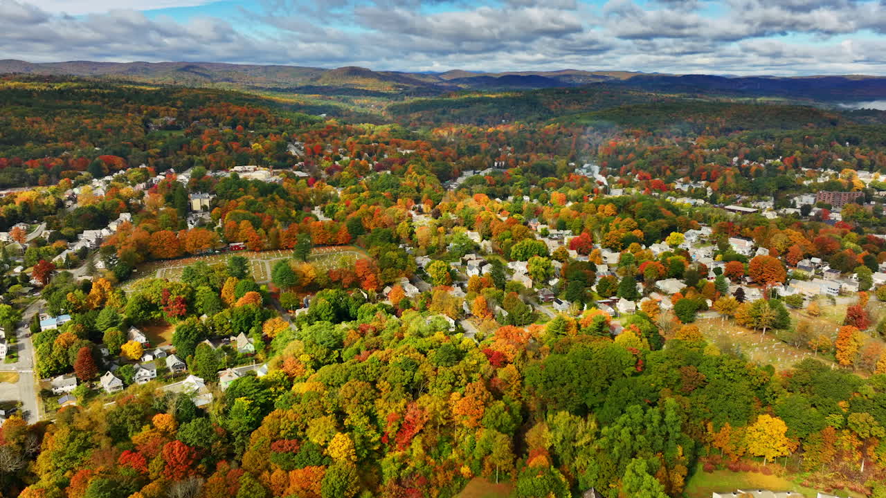 Flying above the colorful trees growing among the cottages. Picturesque mountains under cloudy sky at backdrop. Top view.