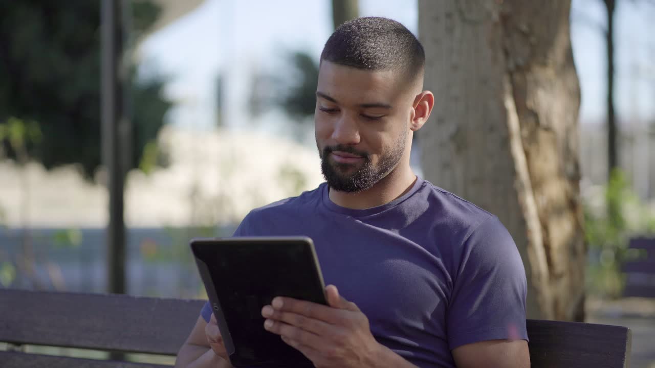 hombre musculoso afroamericano en un banco en el parque escribiendo en una tableta