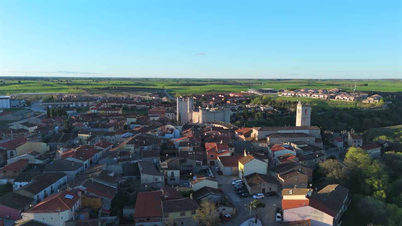 Smooth aerial flight over the Spanish town of Torija, approaching its historic medieval castle with red rooftops and countryside in the background, captured during golden hour with clear skies