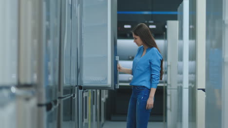 brunette-woman-in-shirt-to-open-the-door-of-the-refrigerator-in-the-appliances-store-and-compare-with-other-models-to-buy-the-new-house