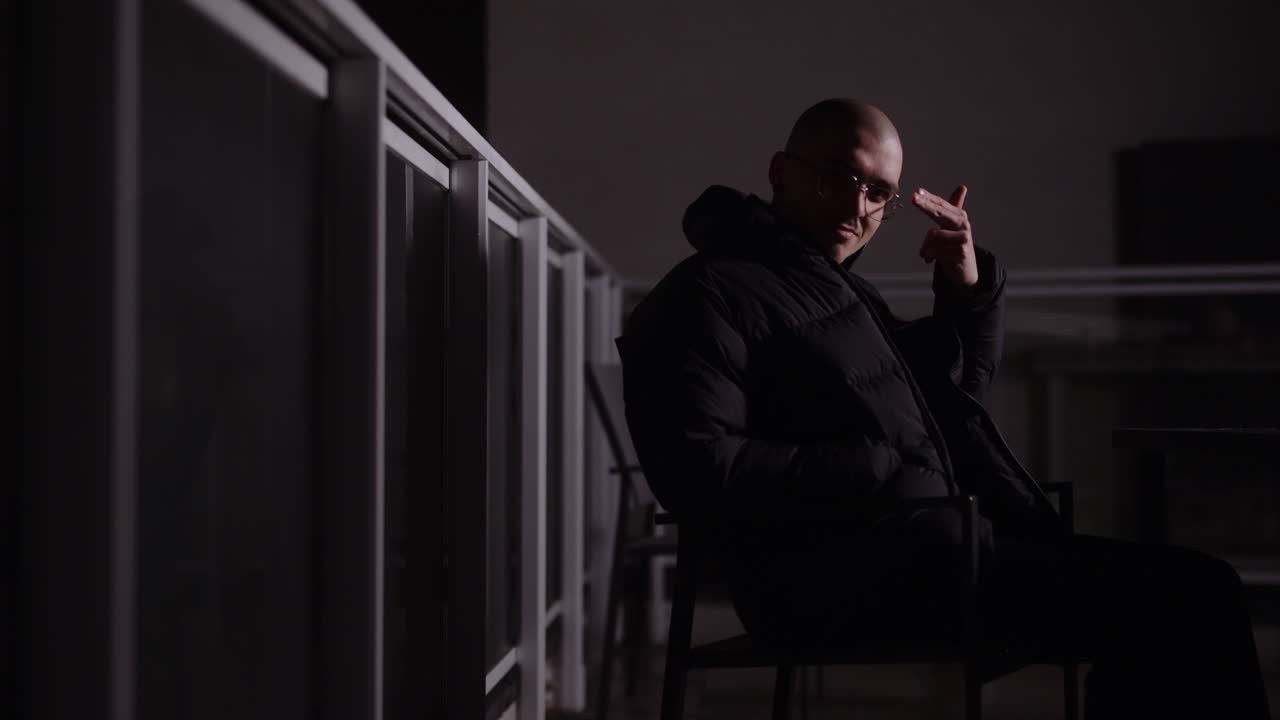 Man in a black puffer jacket sitting on a dark balcony at night