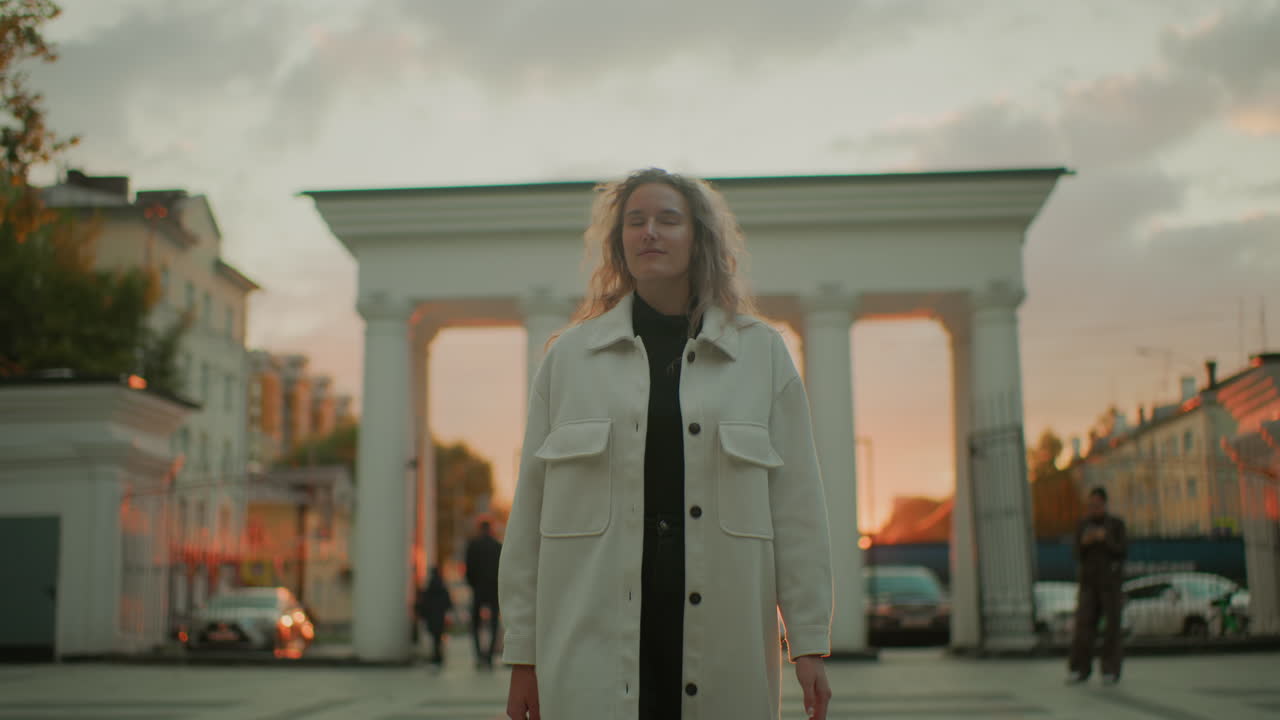 student in long white coat strolling confidently through urban square admiring sunset view as warm golden light highlights her curly hair surrounded by historic buildings people walking and cars moving nearby