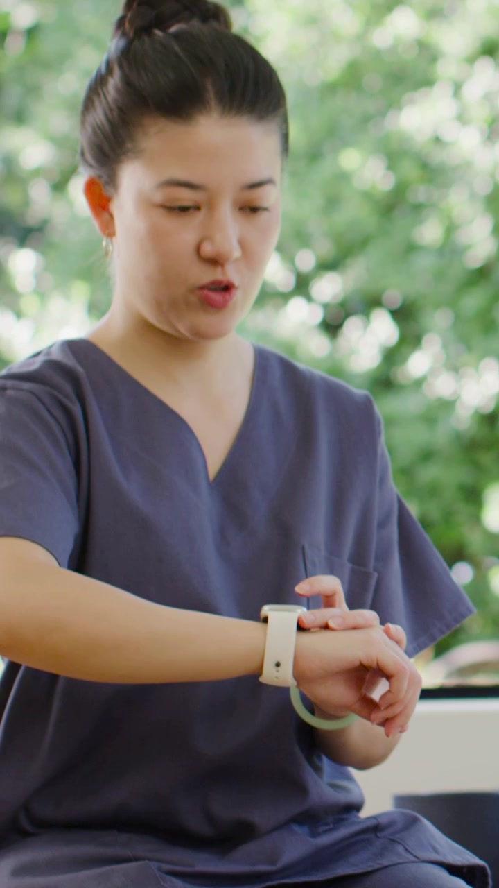 Vertical video: Tapping adult Asian woman in navy scrubs checking white wrist device near window