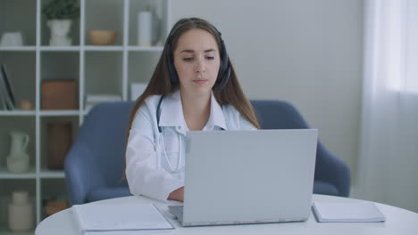 Asistente-Médica-Femenina-Usa-Auriculares-De-Bata-Blanca-Videollamadas-A-Pacientes-Distantes-En-Una-Computadora-Portátil.-Médico-Hablando-Con-El-Cliente-Usando-La-Aplicación-Informática-De-Chat-Virtual.-Concepto-De-Servicios-De-Atención-Médica-Remota-De-Telemedicina.