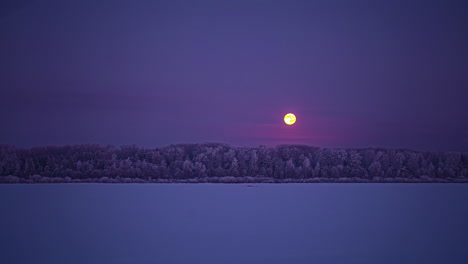 the full moon descends below the atmospheric clouds to the forest beyond a field of snow - nighttime time lapse