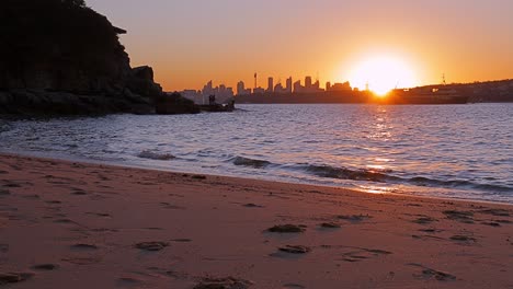 Sydney-CBD-Skyline-silhouetted-by-setting-sun,-beach-foreground,-lapping-waves,-rock-fishers,-ferry---rack-focus,-4K