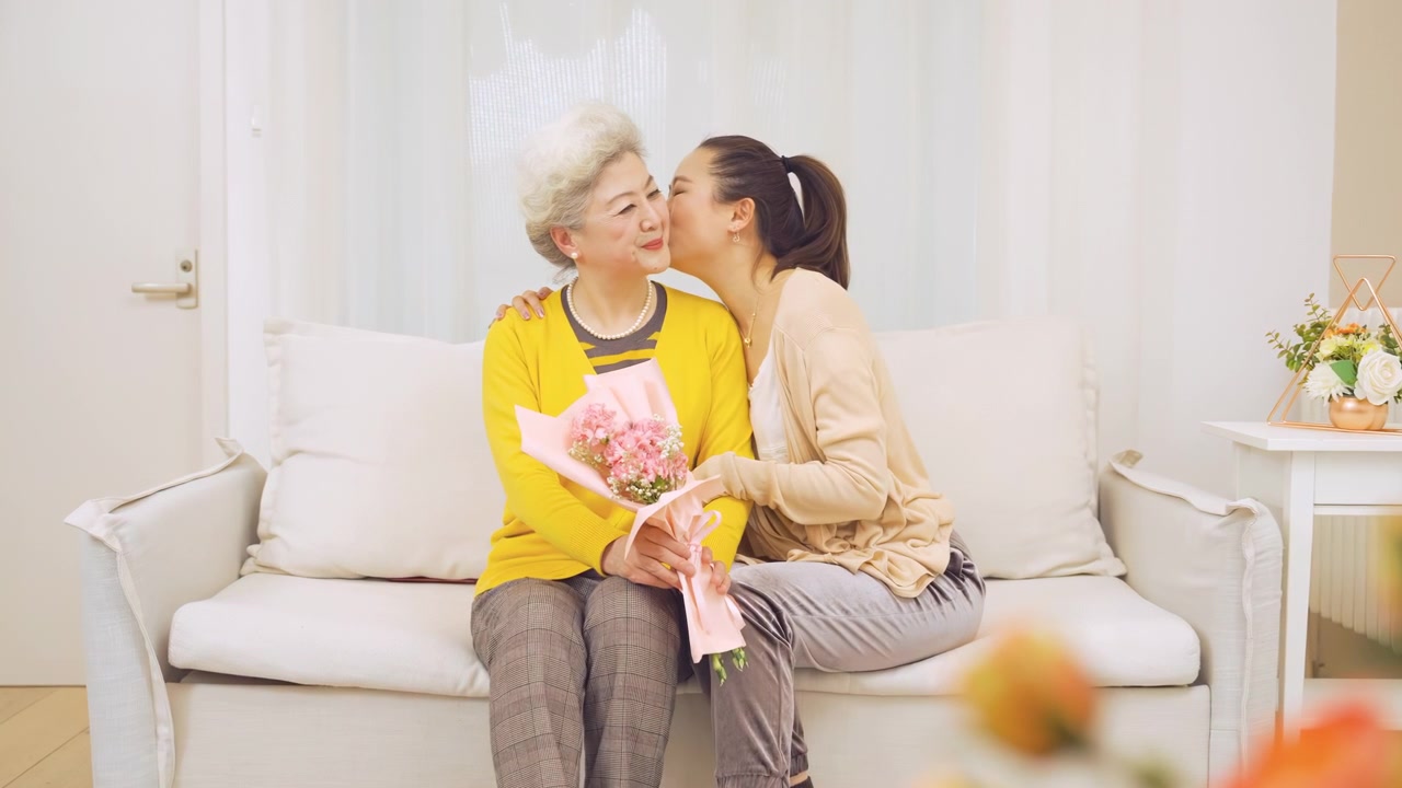 成長した娘が、高齢の母親に花束を贈り、頬にキスをする。母の日を祝う家族のひとときで、退職した母親との関係を大切にする様子。