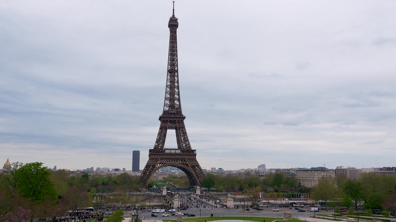 Inquadratura grandangolare centrata della Torre Eiffel che domina lo skyline di Parigi, con strade trafficate, traffico in movimento e pedoni sottostanti. La scena cattura la grandezza del punto di riferimento e la dinamicità della città.