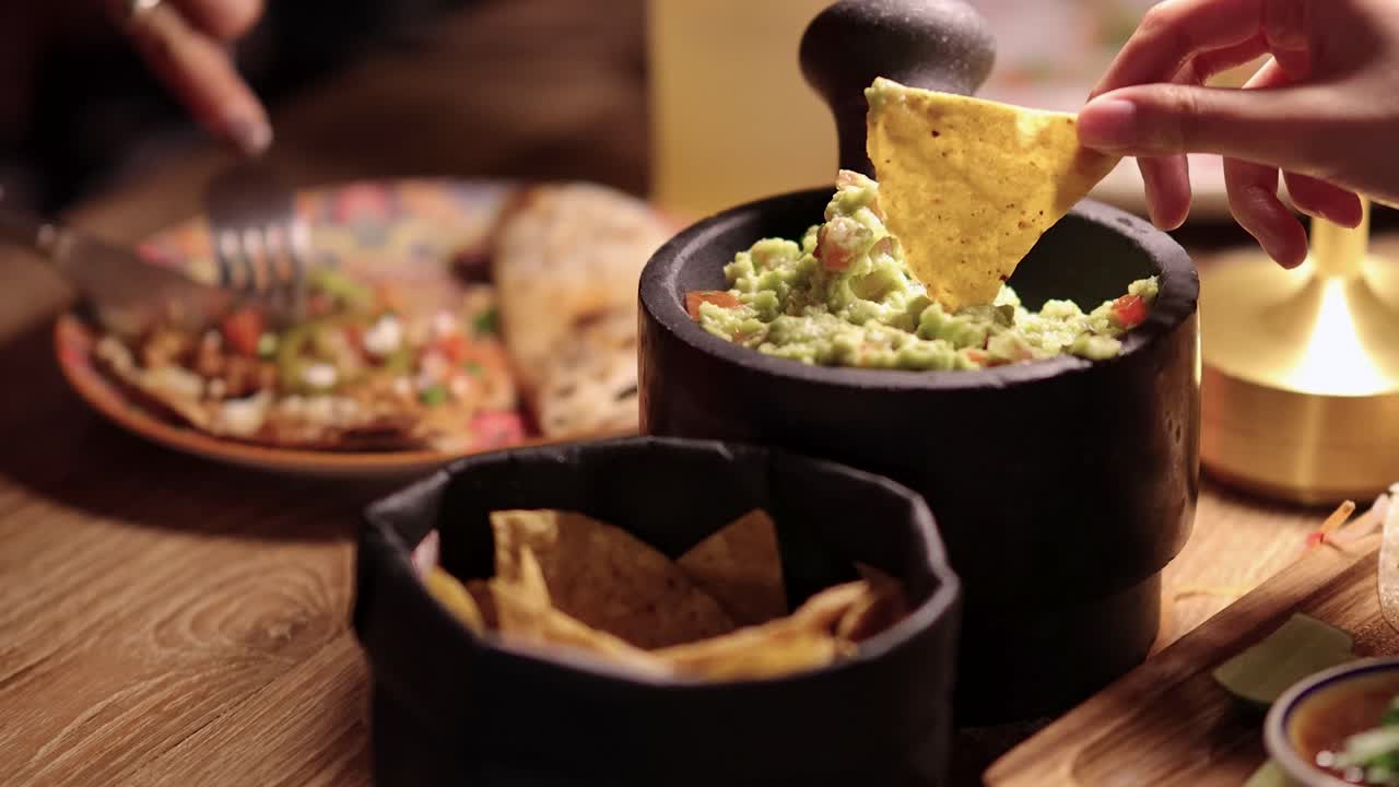 A hand dips tortilla chips into guacamole at a warmly lit restaurant table with grilled quesadilla, salsa, and Mexican cuisine, shot in Bangkok, Thailand