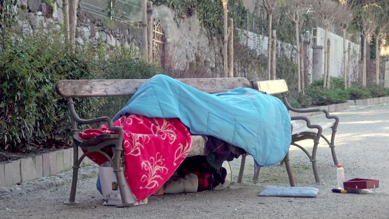 Senzatetto sdraiato sotto coperte e un sacco a pelo su una panchina in una fredda giornata invernale