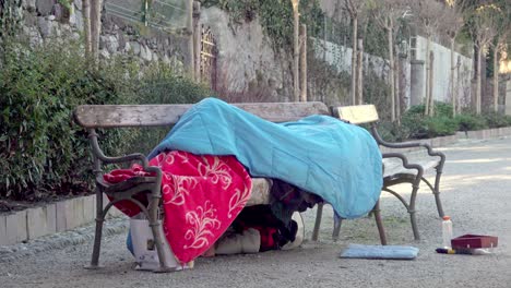 homeless person lying under blankets and a sleeping bag on a bench on a cold winter day