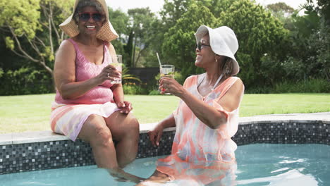 happy senior african american female friends in sunhats sitting in pool with drinks, slow motion