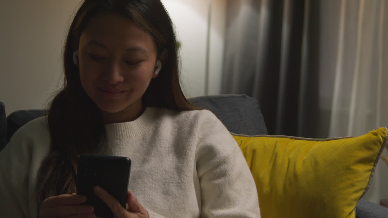 mujer pasando la noche en casa en el sofá usando auriculares inalámbricos y transmisión de música o podcast desde el teléfono móvil 3