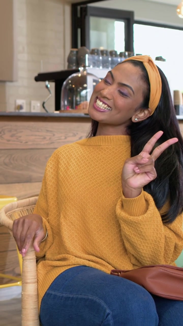 Video verticale: Donna con un maglione color senape che gira a sinistra, toccando un telefono blu scuro in un caffè per un selfie.