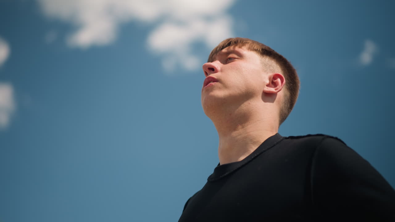 hombre con ropa informal, primer plano de un hombre ajustándose la camisa en el exterior, día informal de verano con un hombre y un cielo azul de fondo, figura masculina relajada vistiendo una camisa negra al aire libre con un ambiente ventoso