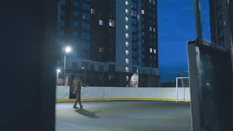 athlete kicks soccer ball toward goal under bright stadium lights on urban sports field, framed by tall residential buildings and a vivid blue night sky