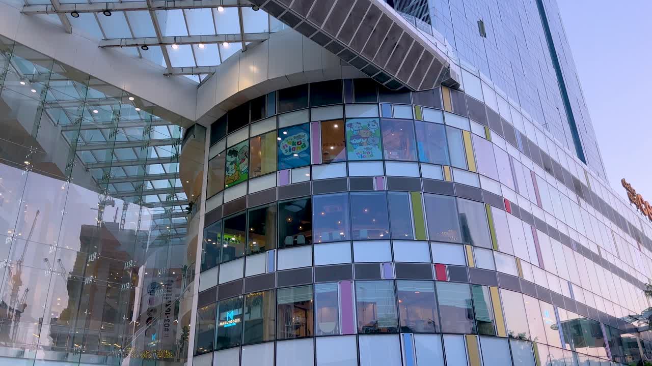 Camera tilts upward along a contemporary glass and steel shopping mall facade in Singapore, capturing architectural details in bright daylight with clear skies