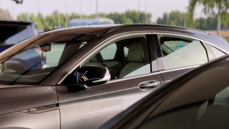 Primo piano di un'auto moderna grigio scuro parcheggiata in un lotto esterno durante il giorno. La luce del sole si riflette sullo specchietto laterale e sui finestrini, con altri veicoli e alberi verdi sfocati sullo sfondo.