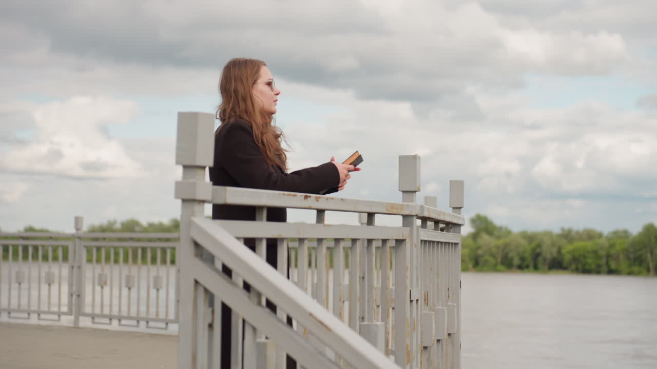 En forretningskvinde i sort frakke og solbriller går selvsikkert med en bog mod et metalgelænder på en åben gangbro under en skyet himmel, der viser beslutsomhed, fokus og elegante bevægelser udendørs