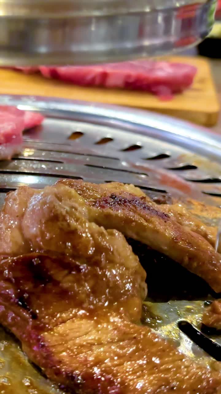 Tampilan dekat daging yang dipanggang di BBQ di restoran Bangkok. Penjepit membalik daging yang mendesis di atas panggangan panas
