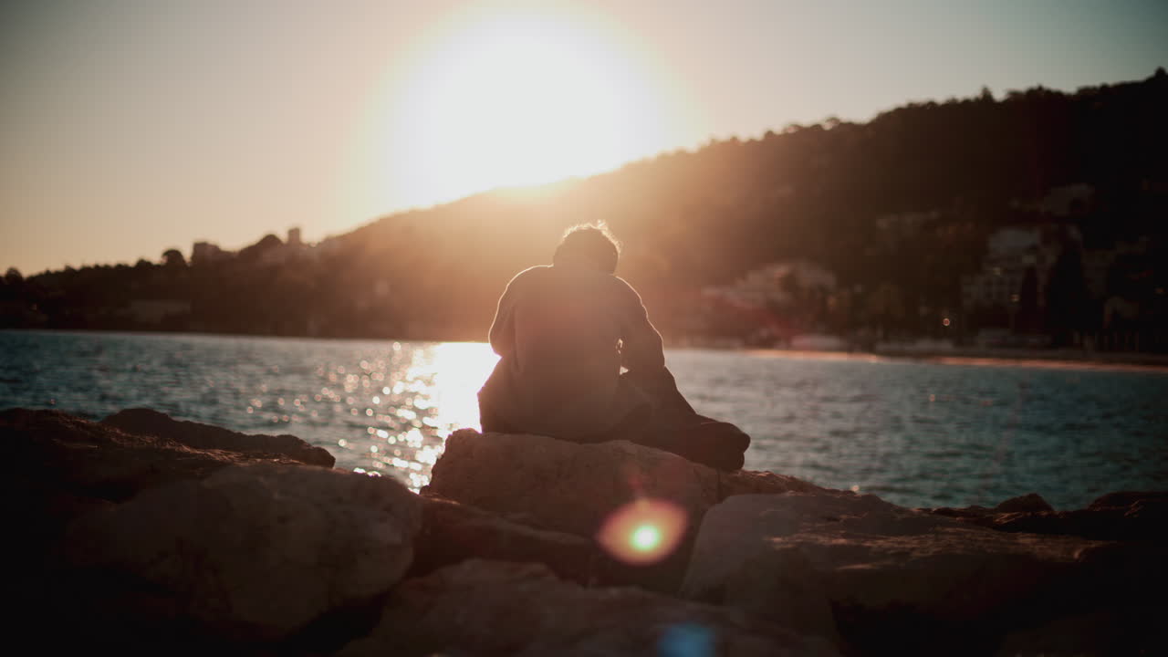 A man sitting alone on coastal rocks, looking down as the sun sets over the sea and hills, creating a warm contemplative mood
